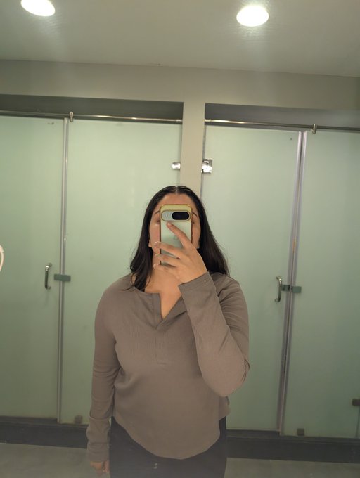 A mirror selfie shows a person with long dark hair wearing a brown long-sleeved henley shirt, holding a smartphone that obscures their face in what appears to be a public restroom.