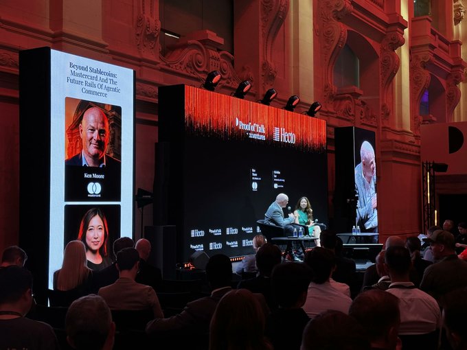 The image depicts a conference stage with several people sitting around a table, engaging in conversation. Some attendees are wearing name tags. The event appears to be focused on crypto topics. There are two prominent speakers on display behind the audience, one of whom is wearing a name tag and the other isn't. Additionally, there's a text posted above the camera that says "There are maybe three crypto conferences left worth attending and @proofoftalk is one of them. Some observations from day 1: 
-@CantonNetwork is the place to build or migrate. They have aspirations to dominate retail payments as well, not just institutional."