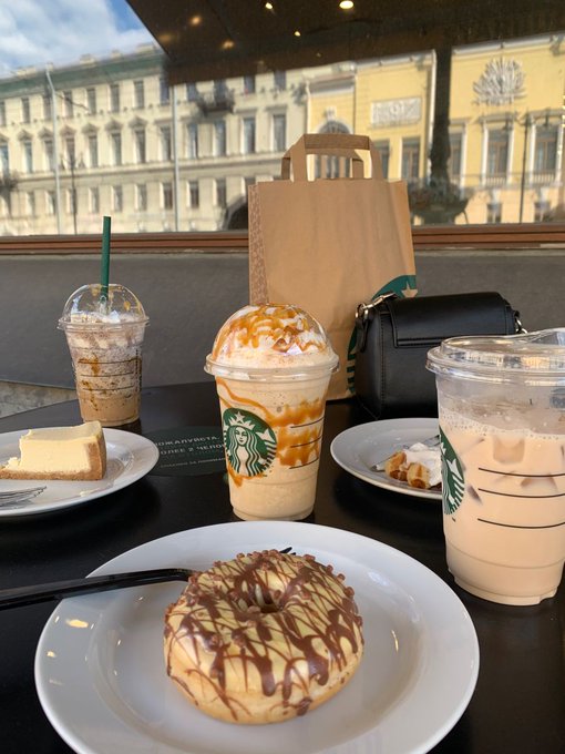 A spread of Starbucks drinks and donuts—including a Frappuccino, a caramel-topped beverage, a latte, a chocolate-glazed donut, and cheesecake—sits on a café table with a paper takeout bag, with European architecture visible through the window. The accompanying post contrasts the always-on, predictable rhythm of Bitcoin's blockchain with the everyday human rituals depicted, like grabbing coffee and a treat at Starbucks.