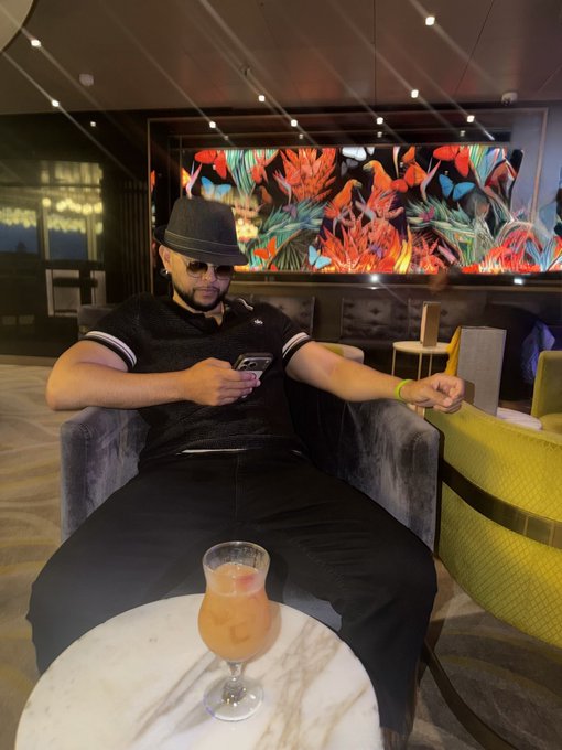 A man wearing a black fedora and sunglasses lounges on a gray chair while looking at his phone, with a cocktail on the table and colorful artwork in the background.