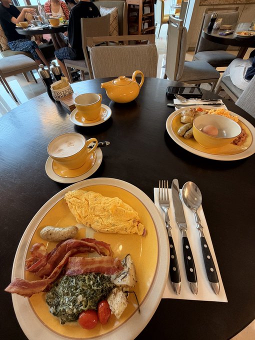 A yellow breakfast plate with eggs, bacon, sausage, and spinach is served alongside a cappuccino and yellow teapot on a dark table, ready for a morning meal.