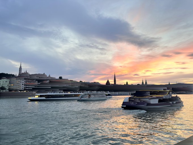 A scenic view of a river at sunset with boats including ones labeled 'rivatours' and 'Wien', with a city skyline and cathedral spires in the background.