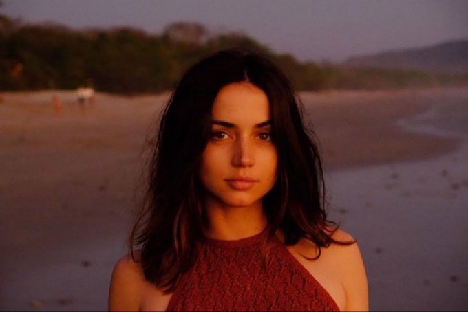A young woman with dark wavy hair and a red halter top stands on a beach at sunset, gazing at the camera.