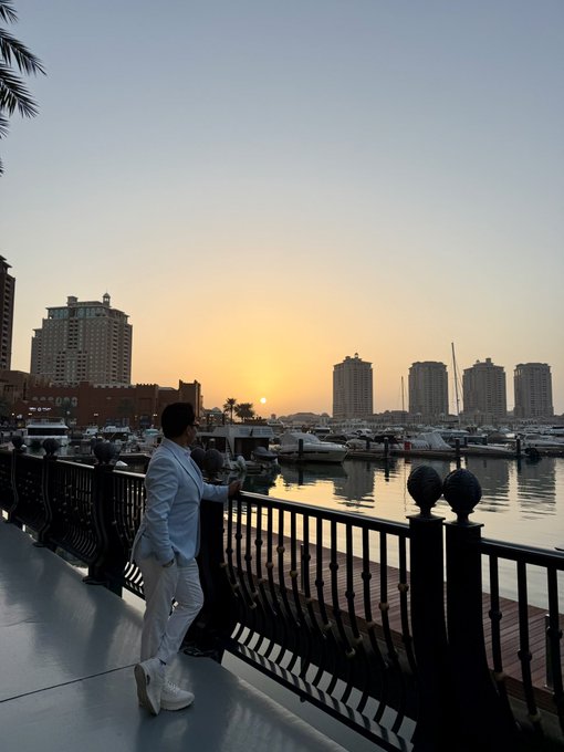 A man in a white suit stands by a black metal railing overlooking a marina filled with boats, with a sunset and city skyline in the background.