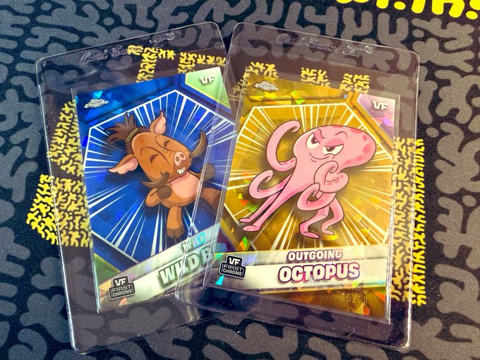 Two VeeFriends "Fight League" sapphire trading cards in protective sleeves are displayed on a patterned surface, featuring a "Wild Boar" with a blue background and an "Outgoing Octopus" with a golden background—both marked "VF" in the corners. The image relates to the post by showcasing the owner's prized 2026 sapphire character cards from a random break, highlighting a milestone moment after previously only obtaining 2025 sapphires.