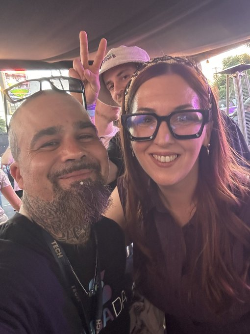 A selfie at what appears to be a festival showing a bearded man with tattoos and lip piercings, a smiling woman with glasses, and another person in the background throwing up a peace sign.