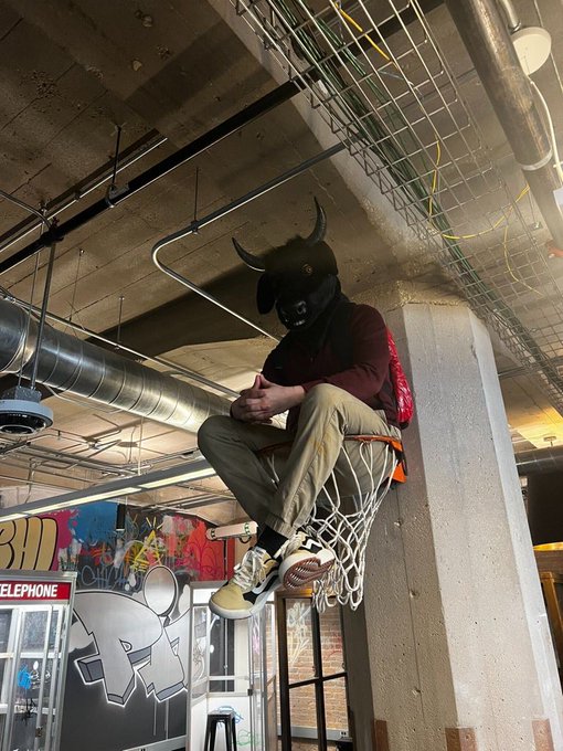 A person wearing a black bull-horned mask, a maroon hoodie, and khaki pants sits on a basketball hoop rim suspended from the ceiling in an industrial-style indoor space.