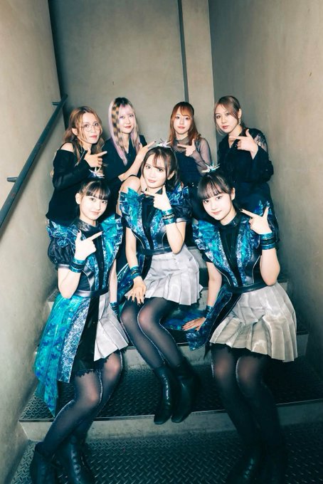 Seven young women pose together on a set of metal stairs, wearing matching teal and black outfits with silver crowns, smiling and making hand gestures at the camera.
