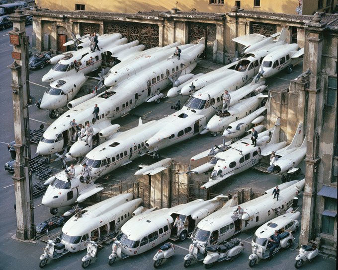An aerial view of numerous small propeller planes and Vespas parked tightly together in what appears to be a hangar or courtyard.