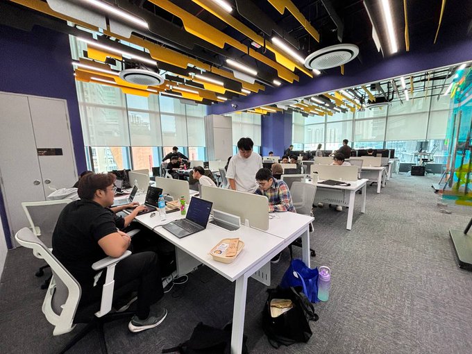 A modern open-plan office with colorful yellow and purple ceiling accents shows several people working at laptops at long white desks.