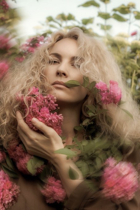 Artistic portrait of a blonde woman surrounded by pink flowers and green foliage, holding flowers close to her face.