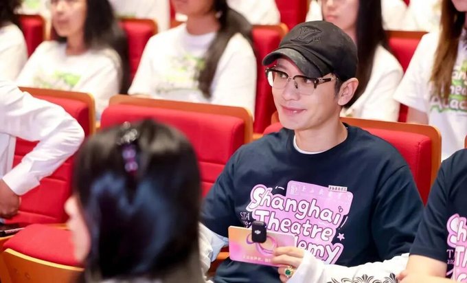 A man wearing a black "Shanghai Theatre Academy" t-shirt, cap, and glasses smiles while holding a microphone, seated among students in a lecture hall, fitting the post about Huang Xiaoming enrolling in Shanghai Theatre Academy's PhD program at age 48.