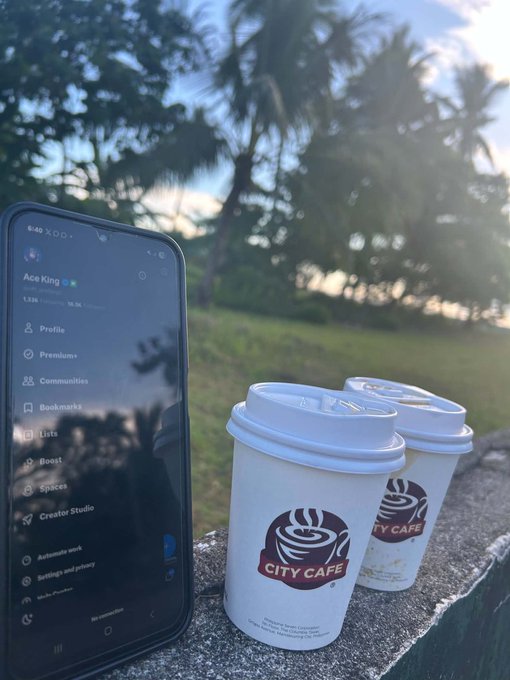 A smartphone displaying the X (Twitter) profile page for "Ace King" sits beside two City Cafe coffee cups on a stone ledge, with palm trees silhouetted against a sunset sky in the background.