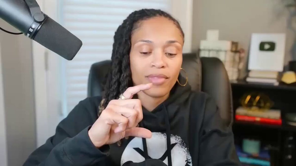 In this image, a woman with long hair wearing a black hoodie holds up her finger as she talks into a microphone, suggesting that she might be discussing an important topic or sharing her thoughts about something. The background features several books, indicating that the setting could be a library or reading area. The caption indicates that the person talking is from BlackRock, and they are part of a pool of institutional investors interested in tokenized U.S. Treasury products. They have reached $2.4 billion in assets, making them the largest single tokenized U.S. Treasury product in terms of market capitalization.