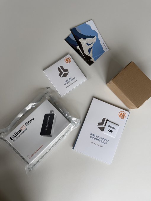 An unboxing photo showing BitBox02 Nova hardware wallet packages including the 'BITCOIN ONLY' hardware wallet, recovery seed cards, tamper-evident security bags, a metallic Bitcoin challenge coin, and a small brown box.