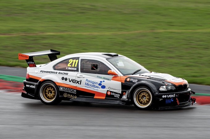 A racing car numbered 211 driven by C. Mackenzie on a wet track, featuring 'POWERDRIVE RACING' branding and sponsor logos including Vexl, Paragon Microfibre, SEEDOR, HALO, and BitcoinBlock on an orange and black livery.