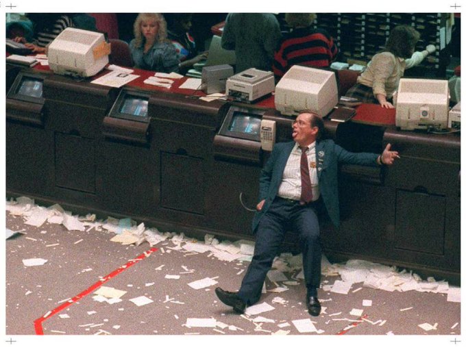 A man in a suit sits amid chaos on the trading floor, surrounded by scattered papers and computer monitors, with people working at desks behind him.