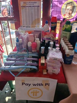 A photo of a merchandise booth selling clothes, bags, and other items with a prominent sign that says 'Pay with Pi' featuring the Pi cryptocurrency logo.