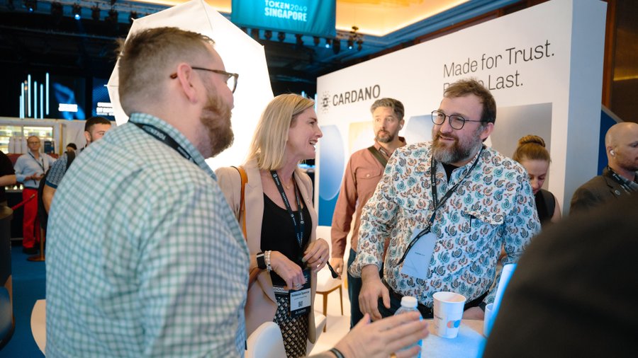 Attendees network at the Cardano exhibition booth at Token2049 Singapore, where visible signage reads "Made for Trust. Built to Last."—capturing a bustling scene of industry professionals in conversation at the event.