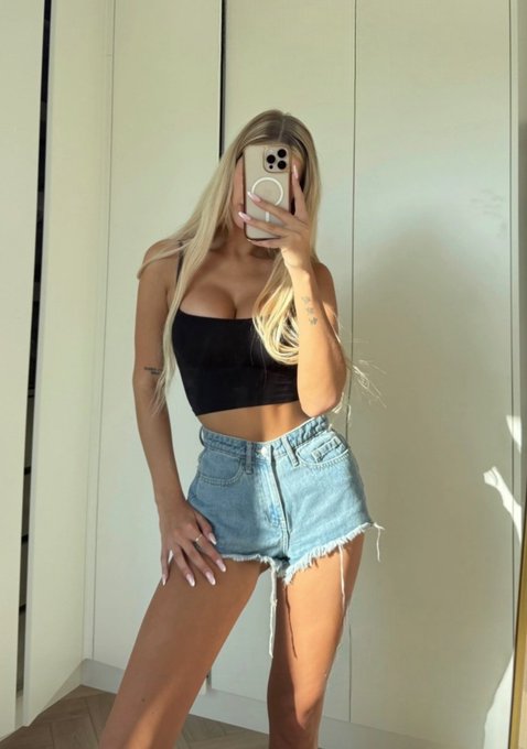 A blonde woman takes a mirror selfie wearing a black crop top and distressed denim shorts.