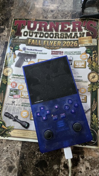 A blue translucent handheld gaming device (resembling an Anbernic RG35XX or similar) rests on top of a "Turner's Outdoorsman Fall Flyer 2026" catalog on a granite countertop.