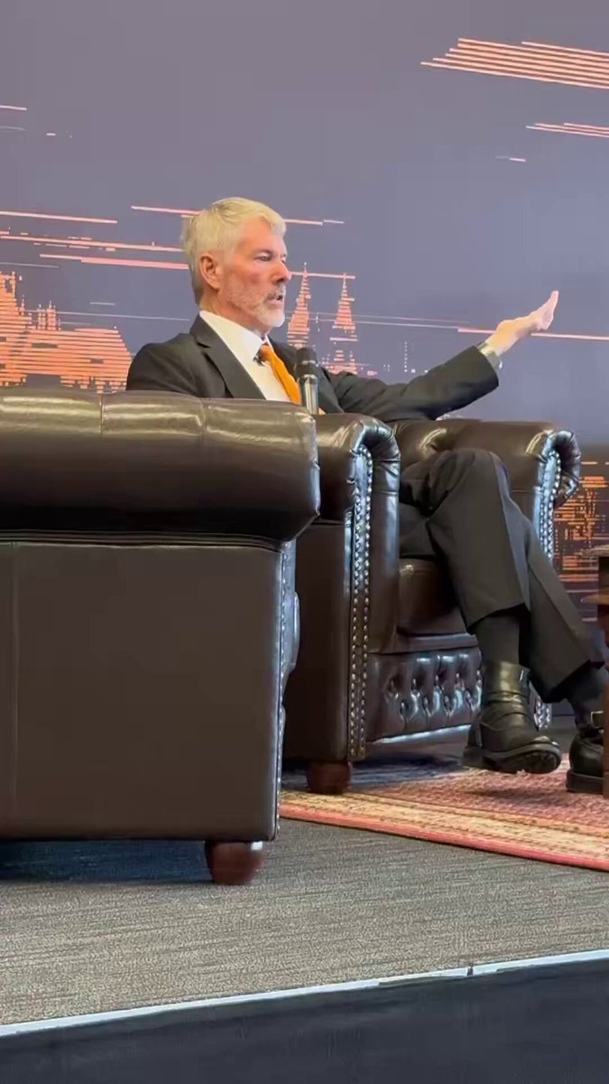 The image features a man sitting in a chair, dressed in a suit and tie, with his hand raised as if he's giving a speech or making a point. The background of the image shows a wall with an orange line running across it, which is likely part of a presentation slide or poster. The caption on the post reads "Saylor: I said not to sell your Bitcoin. I didn't say my company will never sell Bitcoin." This suggests that the man in the suit might be discussing the topic of Bitcoin and its potential impact on businesses.