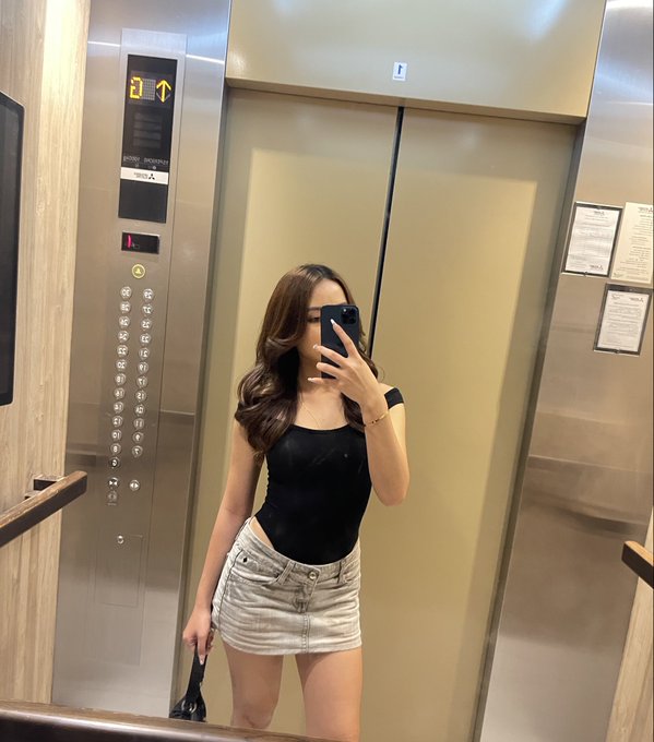 A woman in a black tank top and denim skirt takes a mirror selfie in an elevator, holding a phone that covers her face.