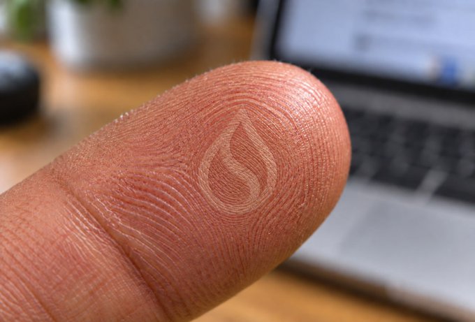 A close-up of a thumbprint with a flame-like swirl pattern in the ridges, photographed in front of a laptop to playfully illustrate the "hands-on" BaseCamp experience on the Sui blockchain.