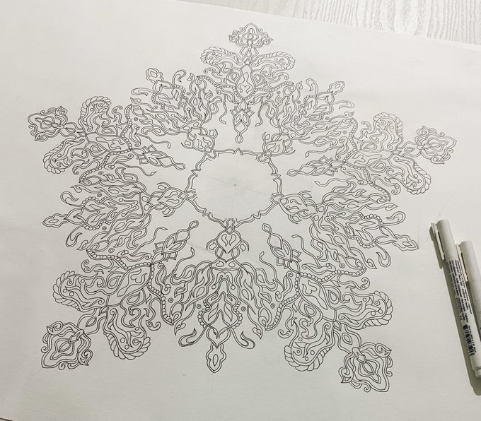 An intricate, symmetrical mandala-style ink drawing on white paper, featuring flowing ornamental patterns with a Micron pen visible at the right edge.