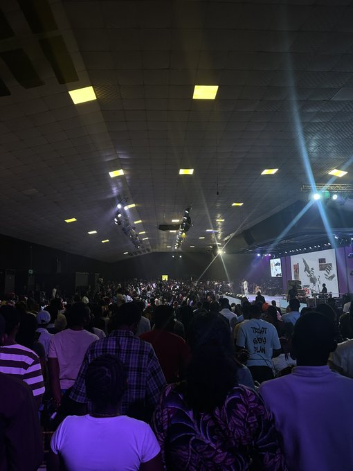 A large crowd attends a dimly lit conference or concert, watching a brightly lit stage with screens at the front of the venue.