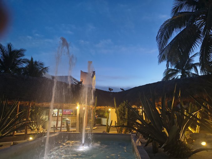 Evening photograph of an outdoor tropical resort area with a water fountain in the foreground, thatched-roof buildings, and palm trees under a dusk sky.
