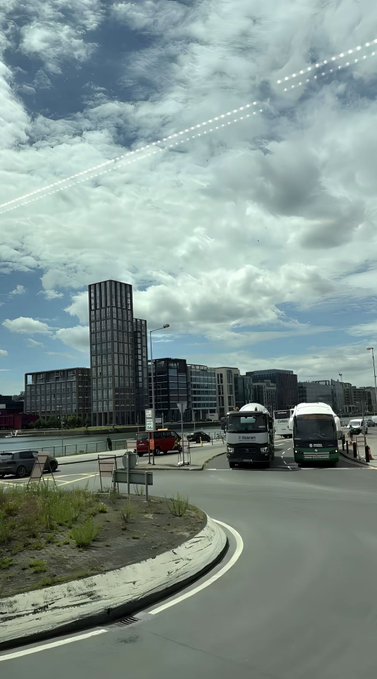 A daytime urban scene with a curving road, parked vans, modern apartment buildings, and a dramatic cloudy sky.