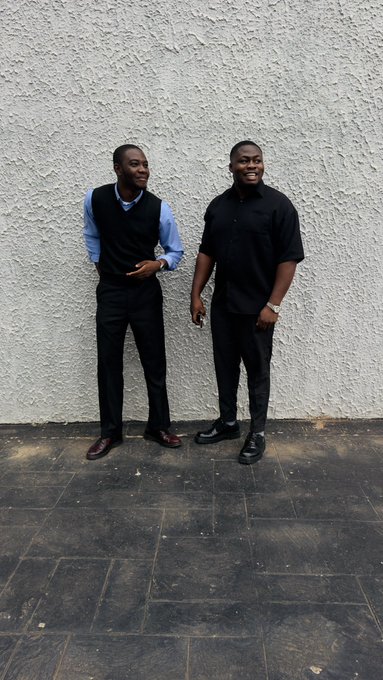 Two well-dressed young men stand smiling against a textured white wall, one in a blue shirt with black vest and the other in an all-black outfit, conveying an air of confident anticipation that complements the post's excitement about ARC mainnet's upcoming launch.