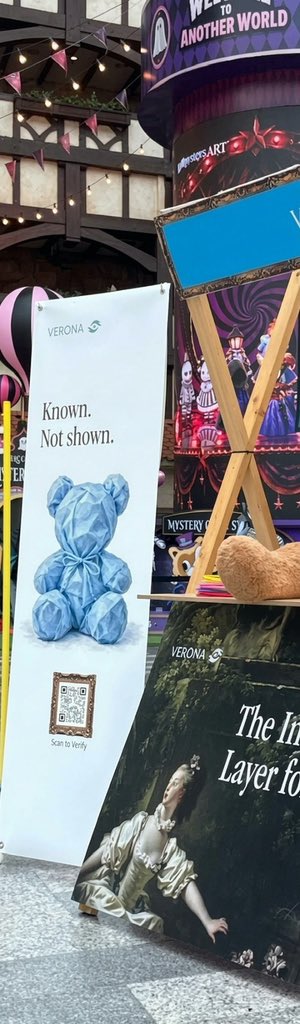 An outdoor art installation or display featuring a Verona banner with a blue geometric bear sculpture reading 'Known. Not shown.' and 'Scan to Verify', set against a backdrop referencing 'MYSTERYS ART' and 'Welcome to Another World'.