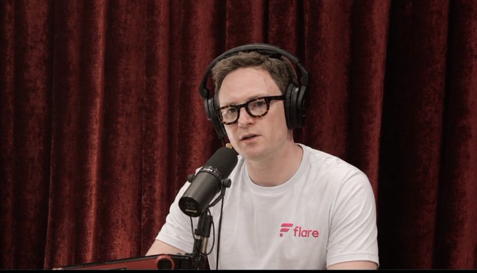 A man wearing glasses, headphones, and a white t-shirt with the 'Flare' logo speaks into a microphone in front of a dark red curtain, appearing to be on a podcast.