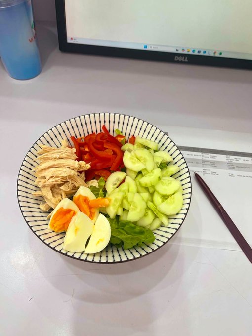 A bowl on a desk contains shredded chicken, sliced red peppers, cucumber, a boiled egg, and lettuce, placed next to chopsticks and a Dell monitor.