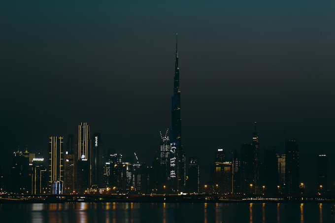 A nighttime cityscape showing the Burj Khalifa prominently illuminated, with Chinese text displayed on a nearby building, reflecting across the water.