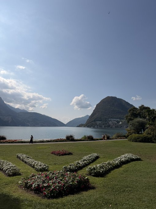 A scenic lakeside garden with a grassy lawn featuring flowerbeds arranged in a decorative pattern, mountains rising across the water under a blue sky with scattered clouds.