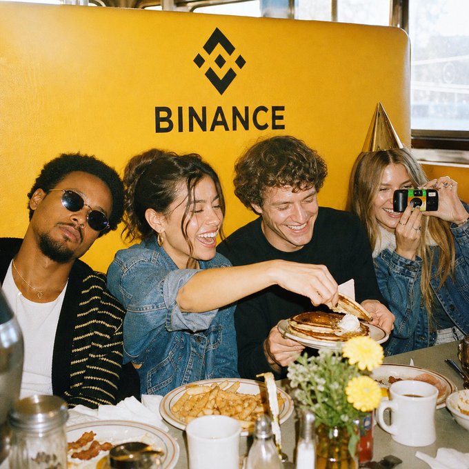 Four friends celebrate a festive occasion at a brunch table in front of a yellow backdrop featuring the Binance logo, with one woman capturing the moment on a small camera as another serves pancakes. The lively scene of shared joy and connection visually reinforces Binance's message about bright moments enjoyed together.