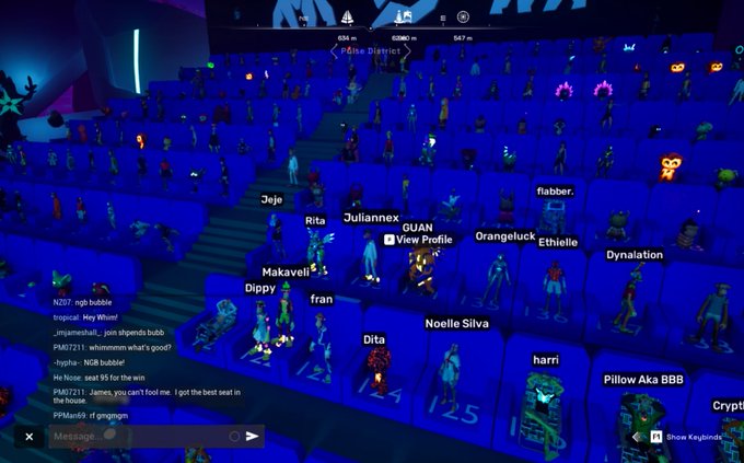 A screenshot from the OthersideMeta virtual world showing an audience of avatars seated in a large blue-lit amphitheater during a film event, with usernames like "Jeje," "Juliannex," "Makaveli," "frank666," "Noelle Silva," "harri," and "Pillow Aka BBB" visible above the avatars, alongside a chat window discussing "NGB bubble" and a "2:5" countdown timer on stage.