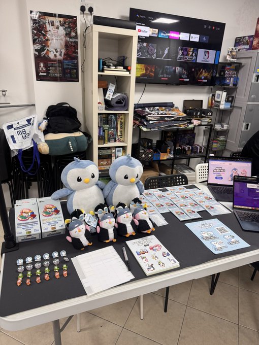 A merchandise table displaying various penguin plush toys, pins, keychains, and printed materials with a laptop on the side.