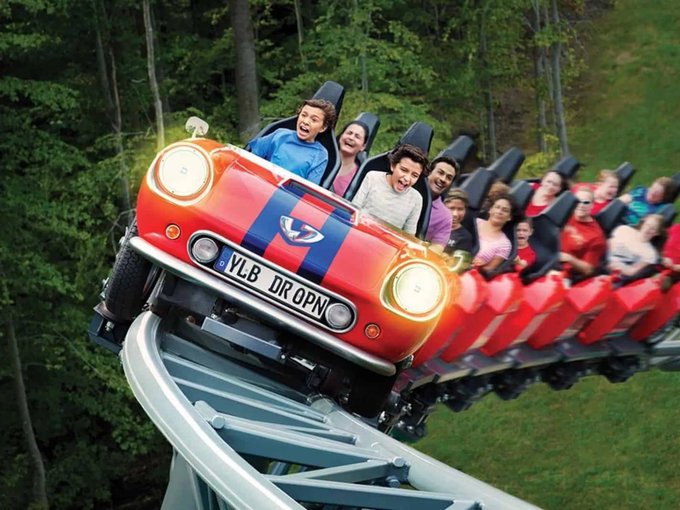 A red roller coaster themed to look like a vintage Volkswagen Beetle with the license plate "YLB DROPN" careens down a track with screaming passengers, perfectly illustrating the post's metaphor about crypto market volatility—bumpy, thrilling, and full of ups and downs that only the most committed riders survive.