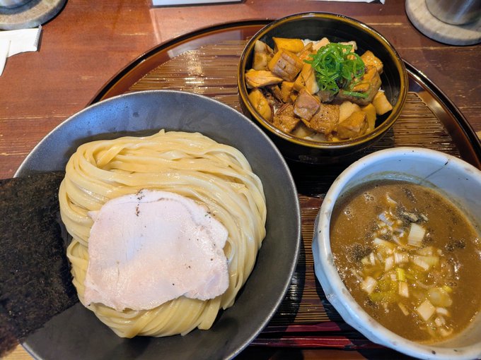 A bowl of thick tsukemen (dipping ramen) noodles topped with a slice of pork and nori, served with a smaller bowl of mushroom and scallion-topped pork chunks and a bowl of dark dipping broth on a wooden tray.