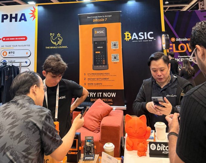 A scene from a trade show booth where representatives from 'BASIC' and 'The Whale Lounge' are demonstrating a Bitcoin payment device to attendees, featuring promotional banners for cryptocurrency products.