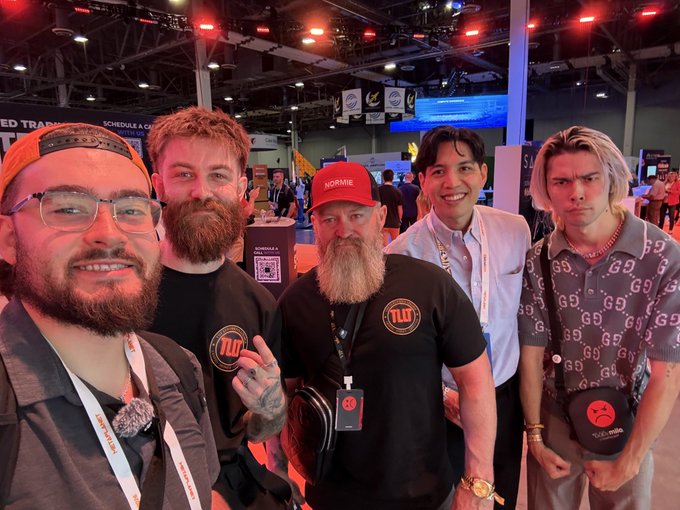 A group of five men pose together at a busy convention, with two wearing 'TLO' branded black shirts and one wearing a red 'NORMIE' cap.