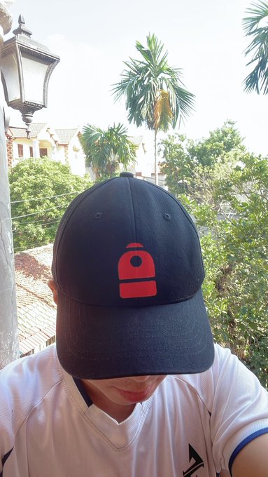 A person wearing a black baseball cap with a red weight/powerlifter logo, photographed against a backdrop of palm trees and a street lamp.