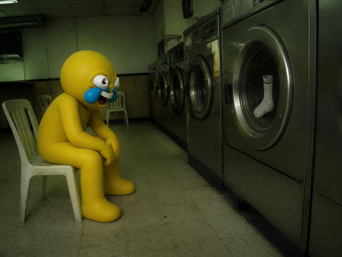 A sad yellow figure with tears sits on a plastic chair inside a dim laundromat, looking toward industrial washing machines where a single white sock rests in the drum. The image humorously illustrates the post's message about patience, as the forlorn character waits for just one sock's wash cycle to finally finish.