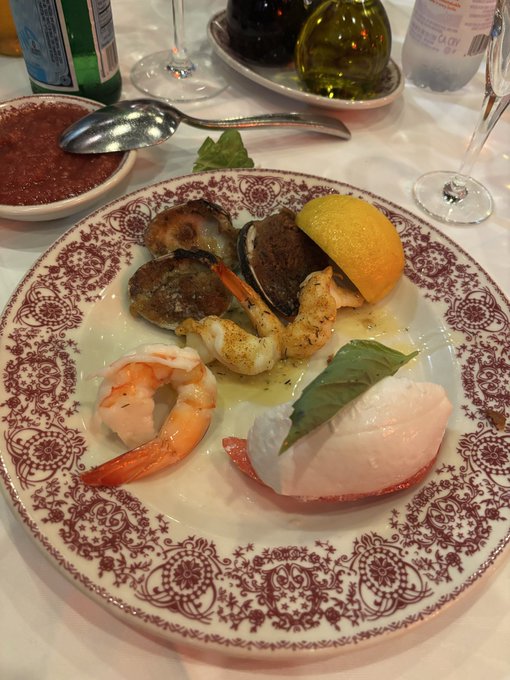 A decorative plate displays a seafood dish featuring shrimp, fish, and what appears to be mushrooms or shellfish, garnished with a lemon wedge and basil leaves, set on a dining table with wine glasses and a bottle in the background.