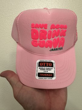A hand holds a pink trucker hat branded with 'SAVE AQU DRINK GUAVA JARritos' and an attached OttO branded 'TRUCKER' hangtag.