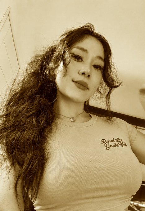 A sepia-toned selfie of a woman with long wavy hair wearing a 'Bored Life Yacht Club' t-shirt and a delicate flower necklace.